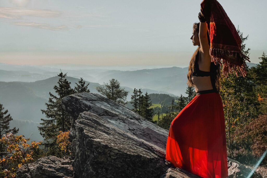 Radikal ungezähmt. Mach dich bereit, Schwester! Kaya Lasika, Gründerin von REWILD.medicine, einem Coaching für Frauen, steht auf einem Felsen, während sie ein rotes Tuch hochhält und in die Weite schaut.